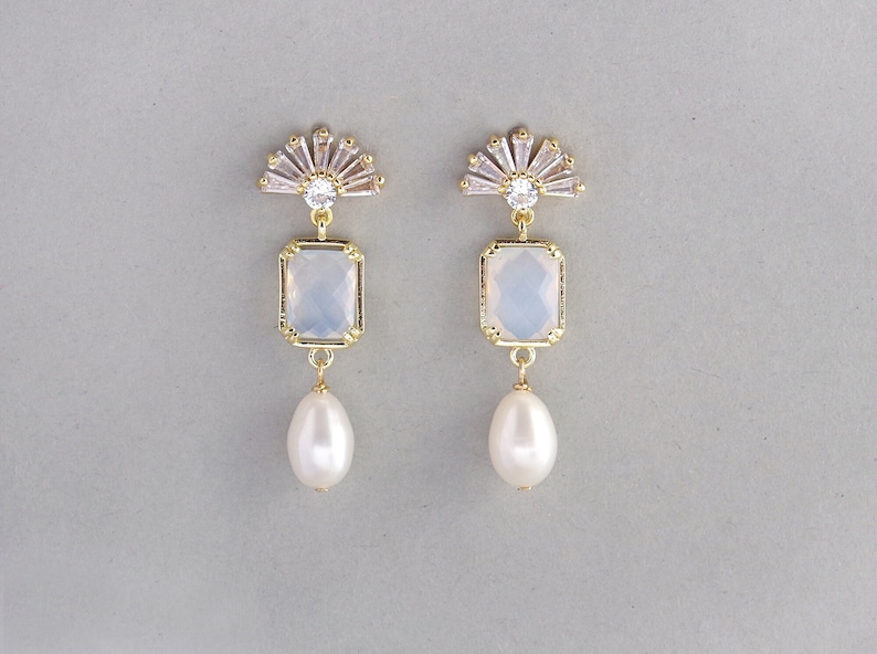 May include: Gold earrings with a white opal-like stone and a pearl drop. The earrings feature a fan-shaped design with clear rhinestones.