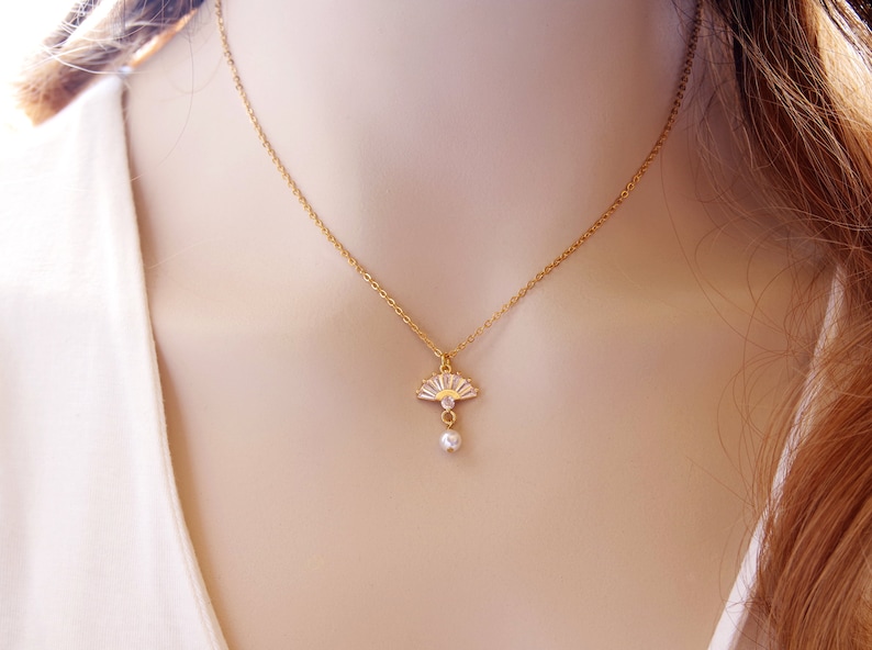 May include: A gold-coloured necklace with a fan-shaped pendant adorned with clear crystals and a small, dangling pearl. The fine chain and pendant design offers a sophisticated aesthetic, suitable for adding a touch of glamour to any ensemble.