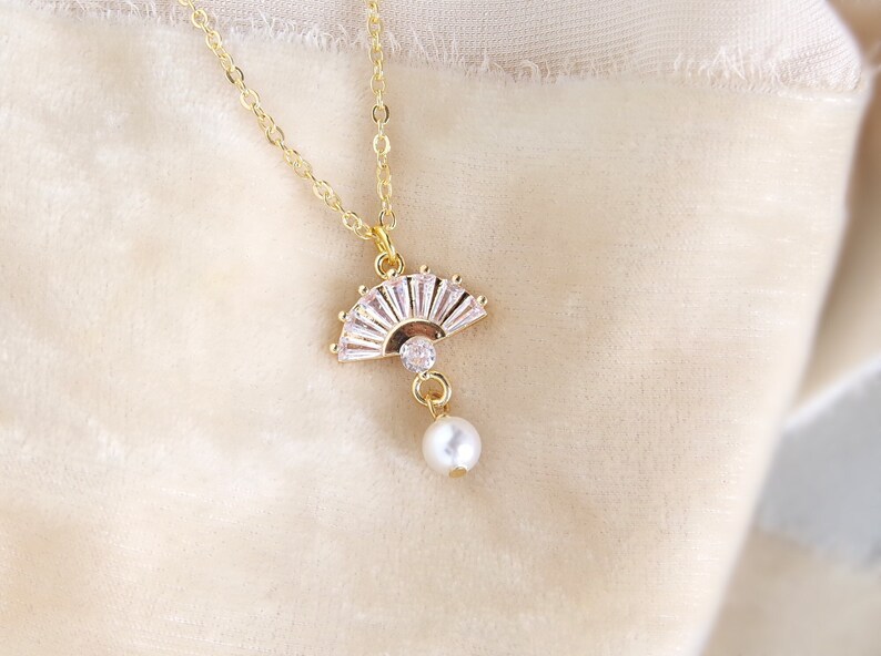 May include: A gold necklace with a fan-shaped pendant set with clear crystals and a central gem. A white pearl dangles below. The necklace is shown against a soft, neutral background. The pendant is a delicate piece of jewellery.