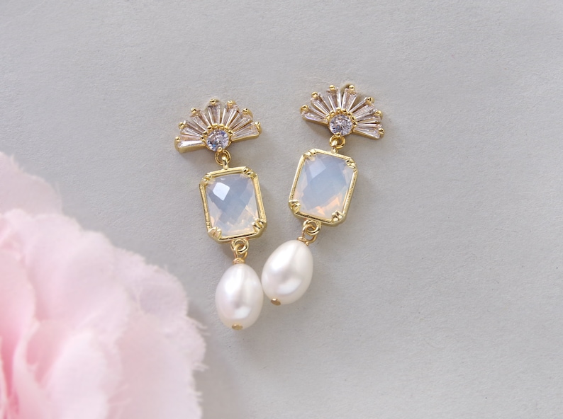 May include: A pair of gold earrings with a white opal-like stone and a pearl dangle. The earrings feature a fan-shaped design with clear rhinestones.
