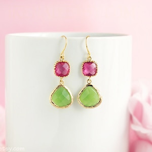 Può includere: Orecchini pendenti con montatura color oro. Ogni orecchino presenta una gemma quadrata color magenta nella parte superiore e una gemma verde a forma di goccia nella parte inferiore. Gli orecchini sono esposti su sfondo bianco.
