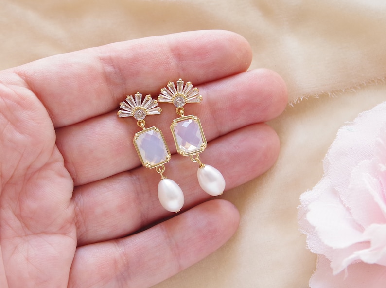 May include: A pair of gold earrings with a clear rectangular stone and a pearl drop. The earrings feature a fan-shaped design with clear rhinestones.