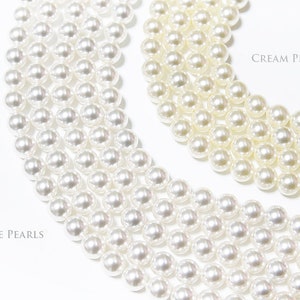 May include: Close-up of white and cream-coloured pearl strands. The image shows multiple strands of pearls, with the text "White Pearls" and "Cream Pearls" labelling the different colours. The pearls are round and have a glossy finish.
