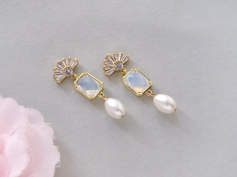 May include: A pair of gold earrings with a white opal-like stone and a pearl drop. The earrings feature a fan-shaped design with clear rhinestones.