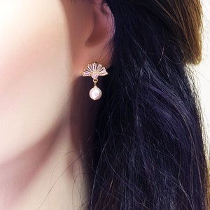 May include: Gold-coloured fan-shaped earrings with pink and clear crystals and a dangling pearl. The earrings are set against a blurred background, highlighting the jewellery's delicate design and elegance.