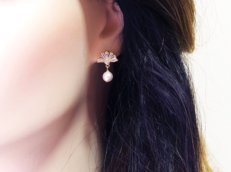 May include: Gold-coloured fan-shaped earrings with pink and clear crystals and a dangling pearl. The earrings are set against a blurred background, highlighting the jewellery's delicate design and elegance.