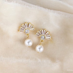 May include: A pair of gold-coloured fan-shaped earrings. Each earring features clear, baguette-cut crystals and a white pearl drop. The earrings are displayed on a soft, cream-coloured fabric.