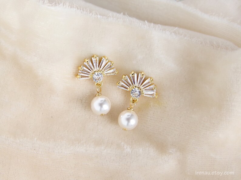 May include: A pair of gold-coloured fan-shaped earrings. Each earring features clear, baguette-cut crystals and a white pearl drop. The earrings are displayed on a soft, cream-coloured fabric.