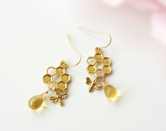 Puede incluir: Un par de pendientes de color dorado con un diseño de panal de abeja con un abalorio de abeja colgante y una cuenta amarilla en forma de lágrima.