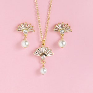 May include: A gold-coloured jewellery set comprising a necklace and earrings. The necklace and earrings feature a fan-shaped design with clear crystals and a dangling pearl. The jewellery is set against a pink background.