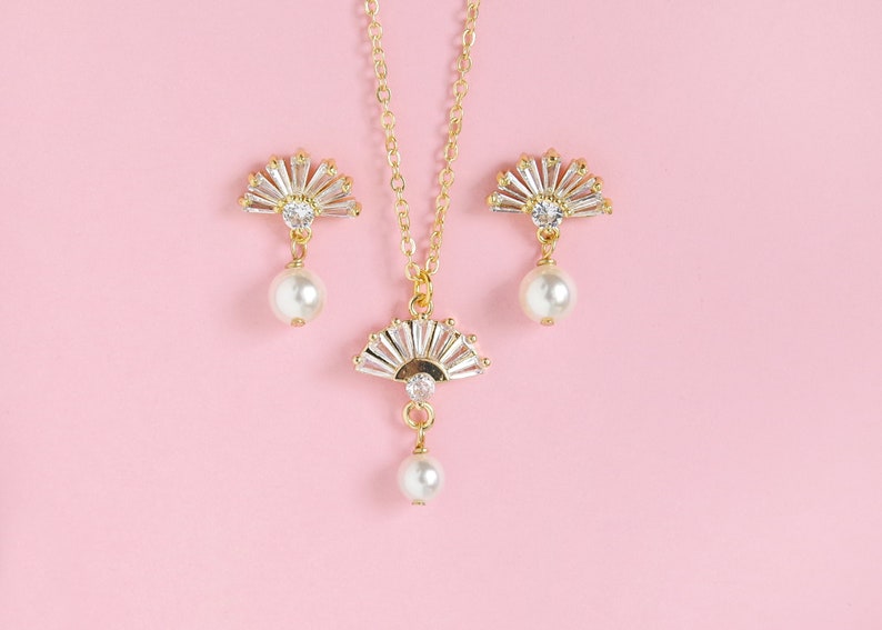 May include: A gold-coloured jewellery set comprising a necklace and earrings. The necklace and earrings feature a fan-shaped design with clear crystals and a dangling pearl. The jewellery is set against a pink background.