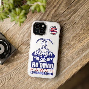 Könnte beinhalten: Eine weiße iPhone-Hülle mit einem blauen und roten Design. Das Design zeigt zwei Hände, die einen Haken halten, mit dem Text "Ho'omau Hawaii Longshoremen" darunter. Ein kleiner Aufkleber mit dem Text "ILWU Local 142" und einer hawaiianischen Flagge befindet sich in der oberen rechten Ecke.