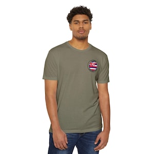 Könnte beinhalten: Olivgrünes T-Shirt mit einem kleinen, kreisförmigen Aufnäher mit der hawaiianischen Flagge und dem weißen Schriftzug "IOWA NOVELTY". Das kurzärmelige Shirt ist aus weichem Material. Die Person trägt eine blaue Jeans.