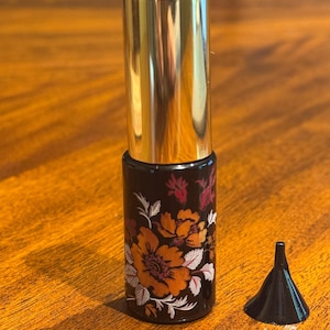 May include: A black perfume bottle with a gold cap and floral design in orange, red, and white. A small black funnel sits next to the bottle. The bottle is cylindrical and appears to be about 4 inches tall.