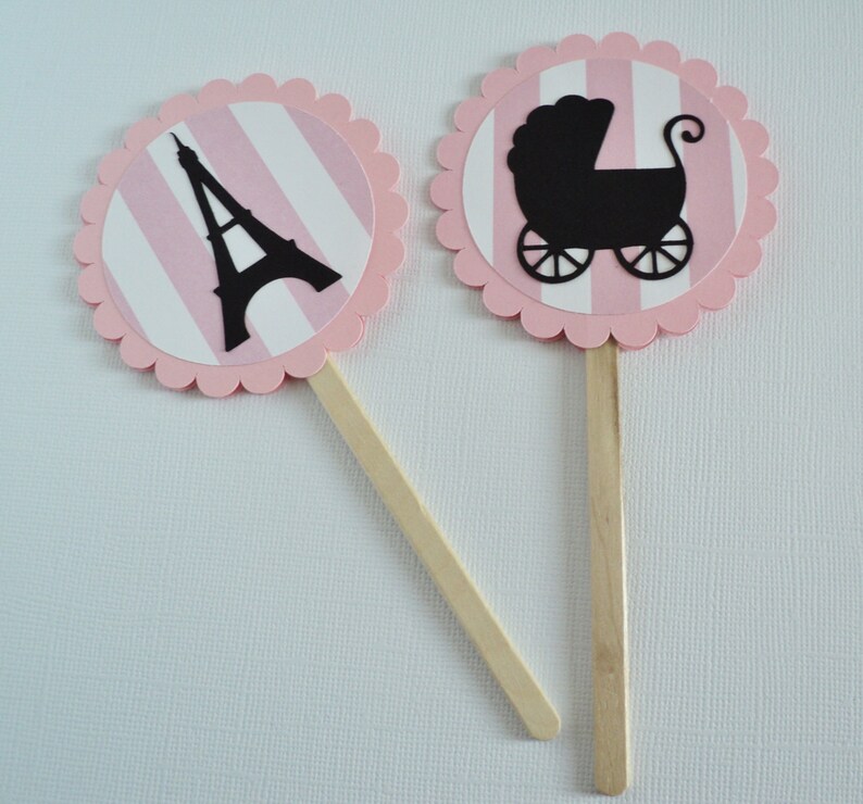 Puede incluir: Dos adornos para cupcakes de rayas rosas y blancas con bordes festoneados. Un adorno presenta una silueta negra de la Torre Eiffel y el otro presenta una silueta negra de un cochecito de beb&eacute;.