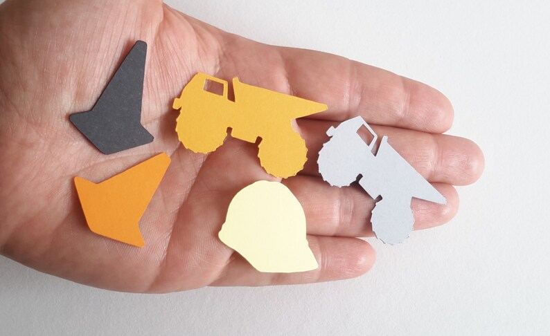 May include: A set of five construction-themed paper cut-outs. The set includes two orange cones, a yellow dumper truck, a grey dumper truck, and a yellow hard hat.