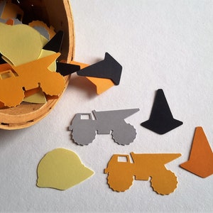 May include: A wooden bucket overflowing with construction-themed paper cutouts. The cutouts include dump trucks, cones, and hard hats in orange, yellow, grey, and black.