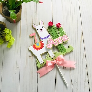 May include: A white llama cake topper with a rainbow saddle and a pink bow. The llama is standing next to a green cactus with pink flowers. The topper says "Anna" in pink letters.