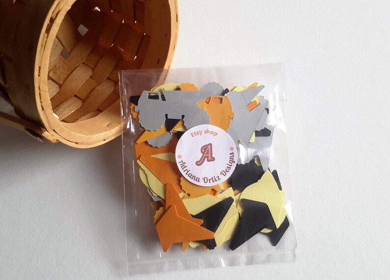 May include: A clear plastic bag filled with confetti featuring construction vehicles, stars, and triangles in black, yellow, and orange. The bag has a sticker with the text "Etsy shop Adriana Ortiz Designs" and a letter "A" in a circle.
