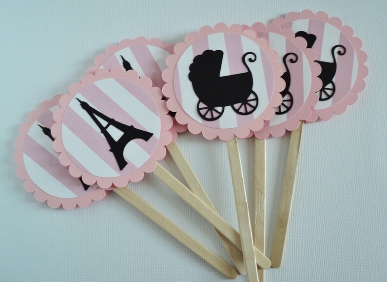 Puede incluir: Toppers para cupcakes de rayas rosas y blancas con siluetas negras de la Torre Eiffel y un cochecito de beb&eacute;. Los toppers est&aacute;n unidos a palos de madera.