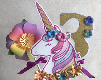 May include: A unicorn cake topper with a gold glitter number 3, rainbow mane, and pastel flowers. The unicorn is white with a gold horn and closed eyes.