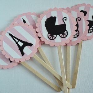 Puede incluir: Toppers para cupcakes de rayas rosas y blancas con siluetas negras de la Torre Eiffel y un cochecito de beb&eacute;. Los toppers est&aacute;n unidos a palos de madera.