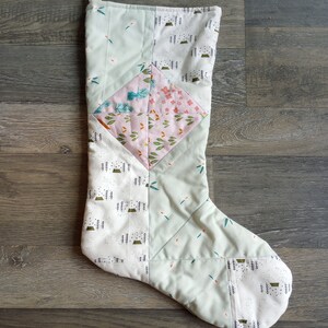 May include: A quilted Christmas stocking with a patchwork design. The stocking features a variety of fabrics in pastel green, white, and pink, with patterns including floral, leaf, and tree motifs. A white loop is attached at the top for hanging.