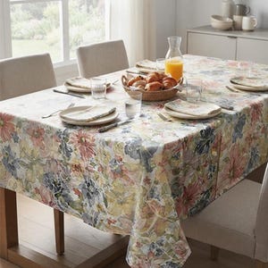 May include: A rectangular tablecloth featuring a watercolor-style floral pattern in shades of pink, blue, yellow, and green. The fabric covers a dining table set with white plates, folded napkins, clear glasses, and a woven basket of croissants.