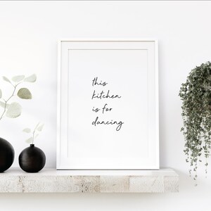 Könnte beinhalten: Ein weißer gerahmter Druck mit dem Text "This kitchen is for dancing" in schwarzer Kursivschrift. Der Druck befindet sich auf einem weißen Regal mit zwei schwarzen runden Vasen und einer Hängepflanze.