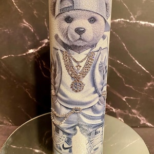 May include: A white glitter tumbler with a cartoon image of a bear wearing a blue and white hoodie, a baseball cap, and a diamond chain. The bear is also wearing sneakers.