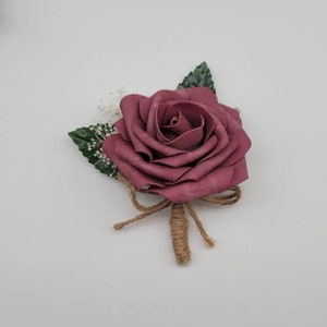 May include: A single burgundy rose with green leaves and a burlap bow. The rose is tied with twine and is a boutonniere.