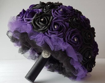 May include: A black and purple bridal bouquet made of silk roses with a black tulle base and a rhinestone brooch.