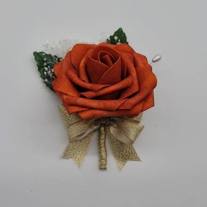May include: A single orange rose with a gold bow and green leaves. The rose is attached to a pin.