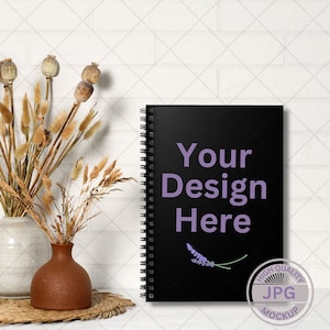 May include: Black spiral bound notebook mockup with the text "Your Design Here" in purple text. A small purple flower is on the cover. The notebook is on a white brick background.