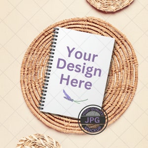 May include: A white spiral bound notebook with the text "Your Design Here" in purple lettering. The notebook is on a woven round placemat with a small lavender flower illustration. A high quality JPG mockup stamp is in the bottom right corner.