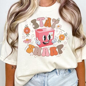 May include: A white t-shirt with a pink and brown graphic design that says "Stay Sharp". The graphic features a pink container with a smiling face and two peace signs. The container is surrounded by flowers and stars.