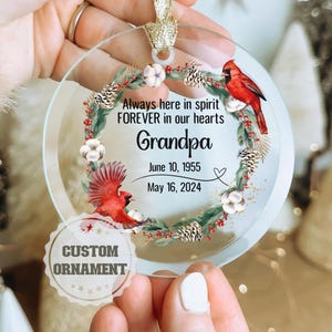 May include: A glass Christmas ornament with a red cardinal, green holly, and white cotton balls. The ornament has the text "Always here in spirit FOREVER in our hearts Grandpa June 10, 1955 May 16, 2024" and "CUSTOM ORNAMENT".
