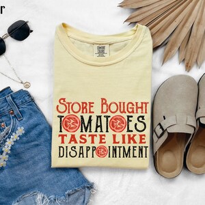 May include: A yellow t-shirt with the text "Store Bought Tomatoes Taste Like Disappointment" printed in black with red tomato slices. The shirt is laying on a white surface with a pair of brown sandals, a pair of sunglasses, a denim skirt, and a gold necklace.