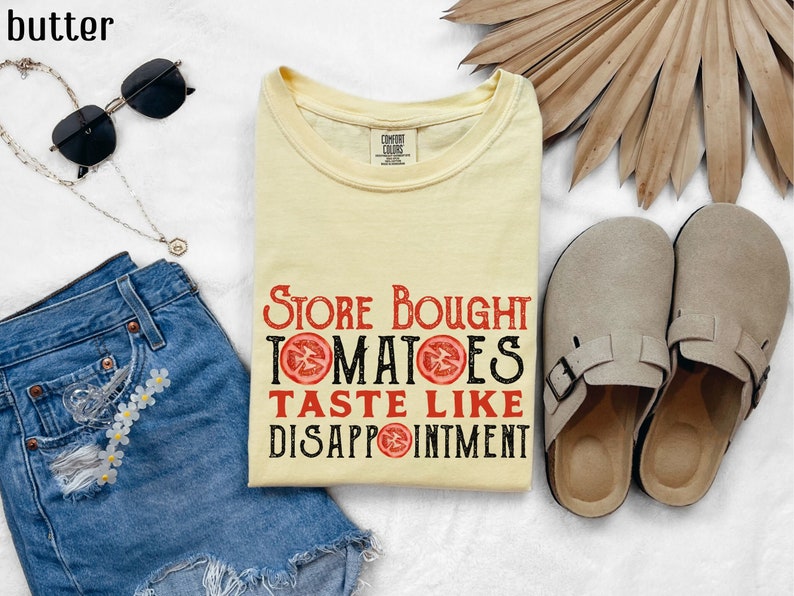 May include: A yellow t-shirt with the text "Store Bought Tomatoes Taste Like Disappointment" printed in black with red tomato slices. The shirt is laying on a white surface with a pair of brown sandals, a pair of sunglasses, a denim skirt, and a gold necklace.