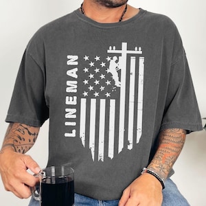May include: A gray t-shirt with a distressed American flag design and the text "LINEMAN" printed vertically on the left side. A silhouette of a lineman working on a power line is visible within the flag design.
