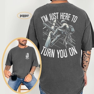 Puede incluir: Una camiseta gris oscuro con un gr&aacute;fico de un lineman escalando un poste de electricidad. El texto "I'm just here to turn you on" est&aacute; impreso en la parte posterior de la camiseta.