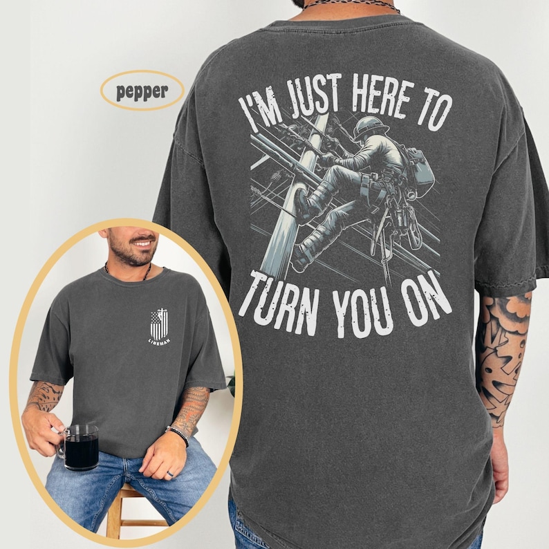 Puede incluir: Una camiseta gris oscuro con un gr&aacute;fico de un lineman escalando un poste de electricidad. El texto "I'm just here to turn you on" est&aacute; impreso en la parte posterior de la camiseta.