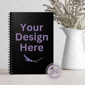 Op de afbeelding: Zwarte spiraalgebonden notitieboek mockup met de tekst "Your Design Here" en een klein lavendeltakje. Het notitieboek ligt op een wit oppervlak met een witte kan en gedroogde lavendel op de achtergrond. De afbeelding heeft een label met een hoogwaardige JPG-mockup.