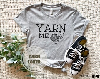 May include: Gray t-shirt with the text "Yarn Me" and a graphic of a ball of yarn with a heart. The text "Yarn Lover" is in a circle below the shirt.