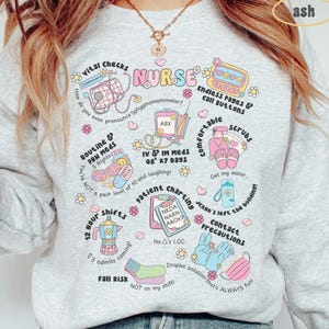 May include: A light grey sweatshirt with a colourful graphic design featuring the word "NURSE" and various illustrations related to nursing, such as a stethoscope, pills, and a thermometer. The text on the sweatshirt includes phrases like "vital checks", "routine & PRN meds", "IV & IM meds", "patient charting", "contact precautions", and "fall risk".