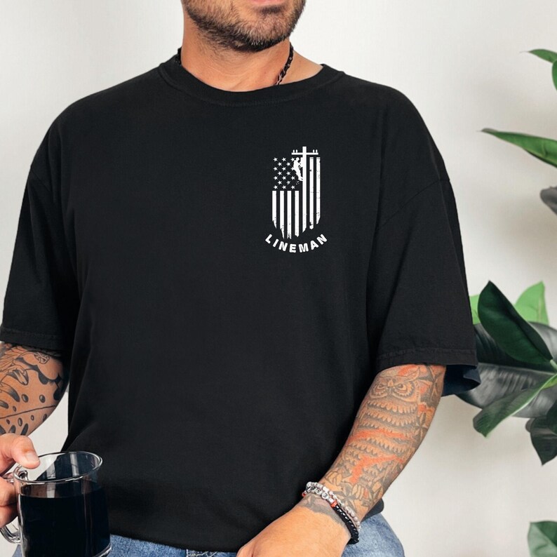 Puede incluir: Camiseta negra con un gr&aacute;fico blanco de un lineman escalando un poste de electricidad frente a una bandera estadounidense. El texto "LINEMAN" est&aacute; debajo del gr&aacute;fico.