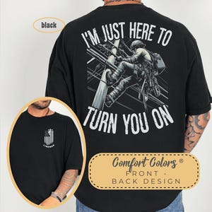 May include: Black t-shirt with a graphic of a lineman on a power pole and the text "I'm just here to turn you on". The front of the shirt has a small American flag with the word "Lineman" underneath.
