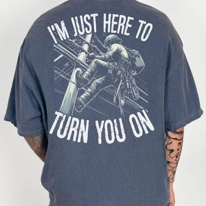 May include: A navy blue t-shirt with a graphic of a lineman climbing a utility pole. The text "I'm just here to turn you on" is printed on the shirt.