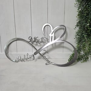 May include: A silver-colored metal wall sign shaped like an infinity symbol with a heart cutout. The sign features the names Ashley and Tony in script, the date 9.16.15, and the number 11 integrated into the design with small heart accents.