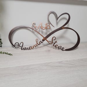 May include: A brown metal infinity symbol with a heart and the names "Amanda" and "Steve" cut out. The date "5-4-18" is above the infinity symbol. The piece is on a light gray wooden surface.